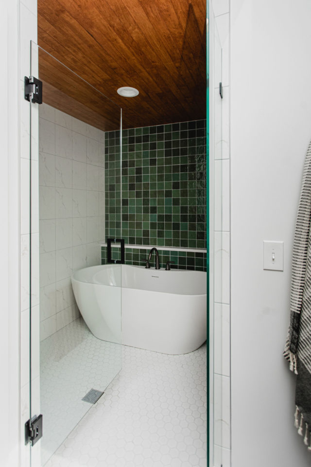 a photo os a custom bathroom with emerald tile on one wall and a freestanding tub with beautiful white hexagon tile on the floor