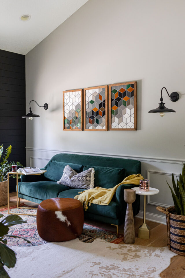 a photo of a custom sitting room with three pieces of art on the wall featuring tile from mercury mosaics, with a pair of wall scandinavian-inspired sconces, a green emerald velvet couch and a cow pouf in hide in brown and two statement layered rugs 