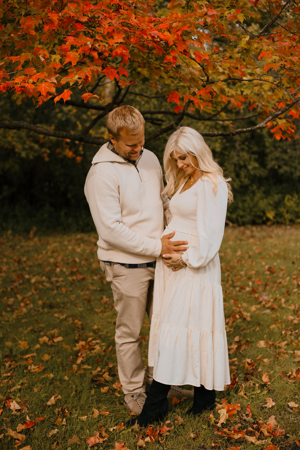 Noah and Anya posing together in the park with beautiful fall color leaves in the trees, Noah has his hand on Anya stomach, who's 8 months pregnant with Solange