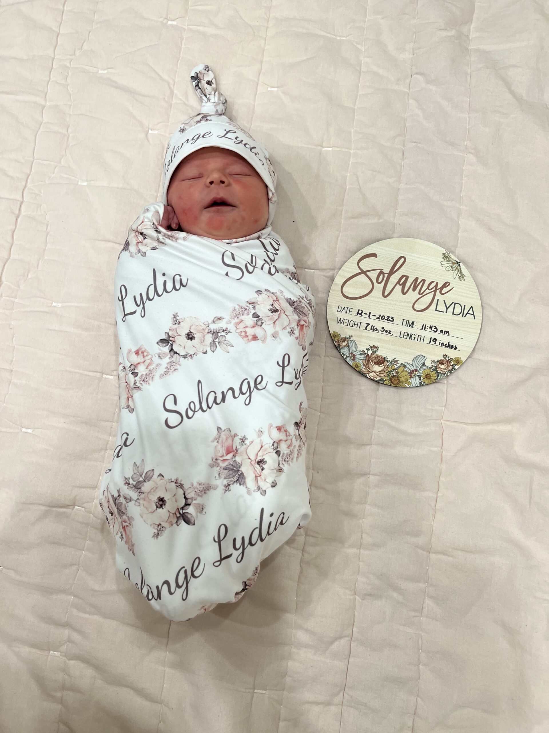 Solange Lydia just a week or two after she was born (VBAC) she's wearing a matching white swaddle and hat, monogrammed with Solange Lydia and flowers and a plaque laying next to her with her name, birth date, weight, heigh, and length written on it.