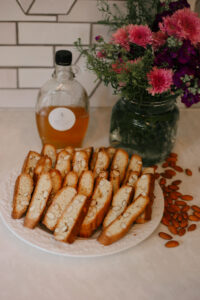 a plate full of freshly baked almond biscotti with a jar of honey and a bouquet of flowers in the background