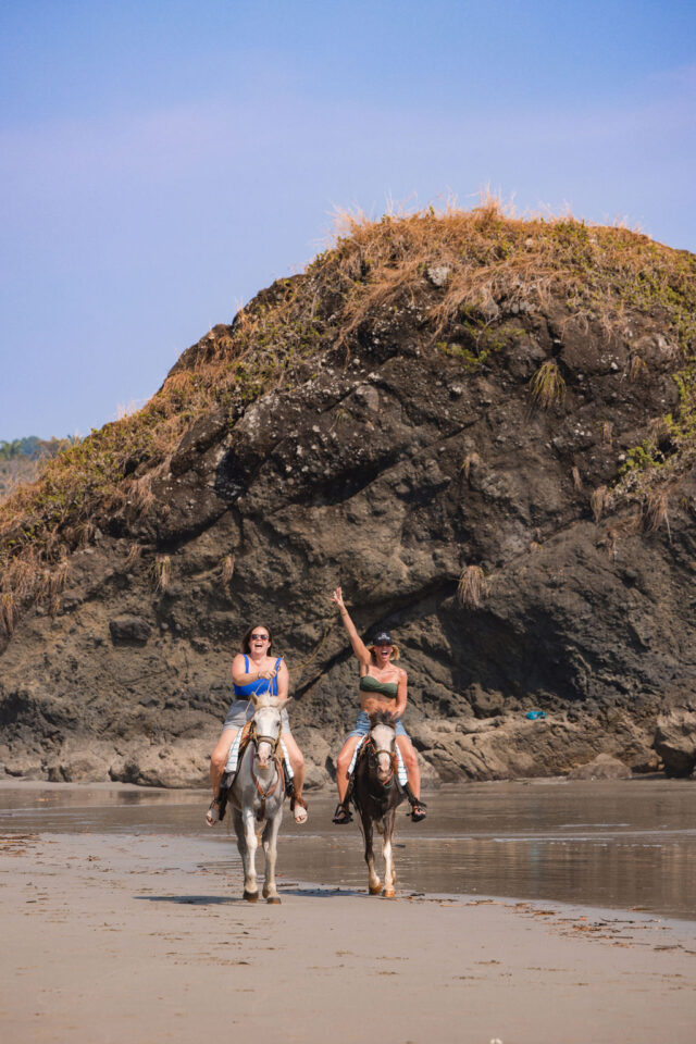 a photo of two female contractors who are excited, riding horses on the beach in Costa Rica