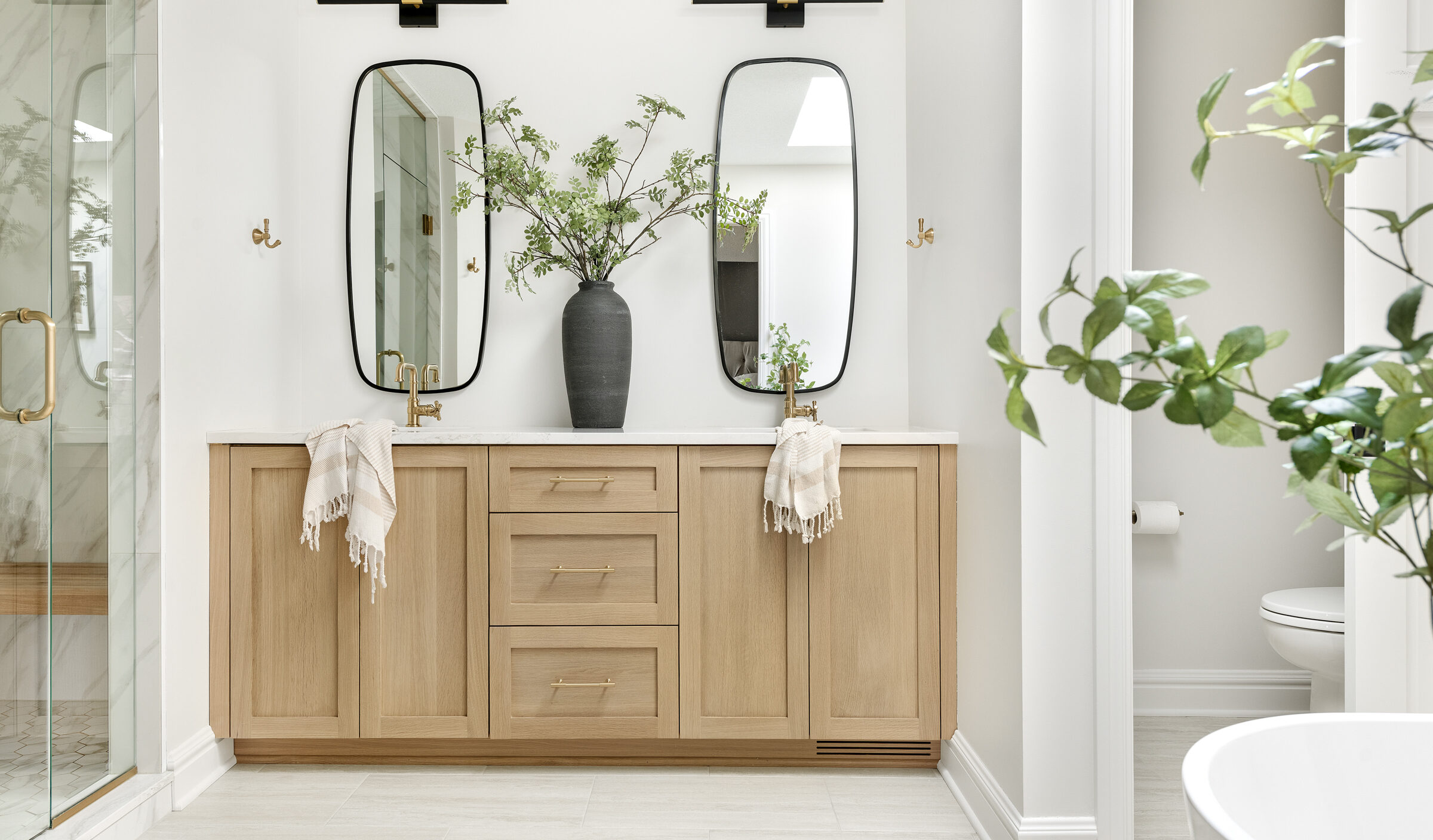 a photo of a primary bathroom in Minnesota designed and built by construction2style, it features a shower, beautiful double vanity in rift sawn white oak, gold hardware, two Rochester Accent Mirrors placed vertically under LED vanity lights and a free standing tub